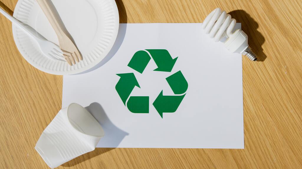 Símbolo da reciclagem e sustentabilidade impresso em uma folha. Ao redor um copo, um garfo, um pratinho de plástico e uma lâmpada.  