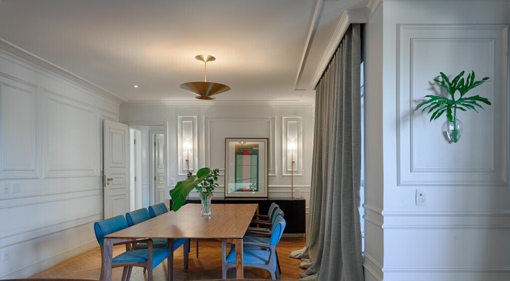 Imagem de uma sala de jantar com decoração em tons de branco, cinza, marrom e azul. Nela há uma mesa de jantar em madeira com cadeiras azuis, cortina cinza e paredes brancas com boiseries brancos, simbolizando o processo de acabamento em obras.