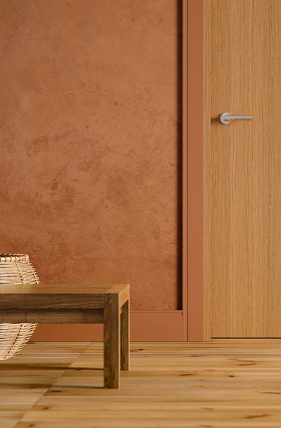 Imagem de uma porta e parede em tons terrosos. Nela também há um banco de madeira com um vaso atrás e sócalos e rosetas simbolizando o processo de acabamento de uam obra.