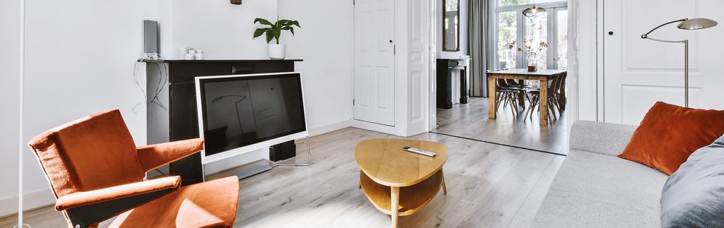 Sala de TV com decoração contemporânea. Suas paredes são brancas, revestimento de piso claro, mesa de centro em madeira, sofá cinza, poltrona e almofada em laranja-escuro.