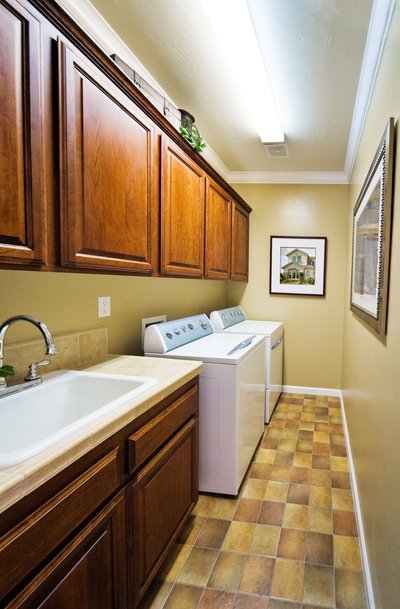 Área de serviço pequena com armários de madeira suspensos, máquinas de lavar e secar, e quadros na parede.