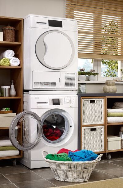 Área de serviço pequena com armário na vertical no lado esquerdo da foto, máquina de secar em cima de uma máquina de lavar, armário de chão no lado direito e uma cesta de roupas em frente as máquinas.