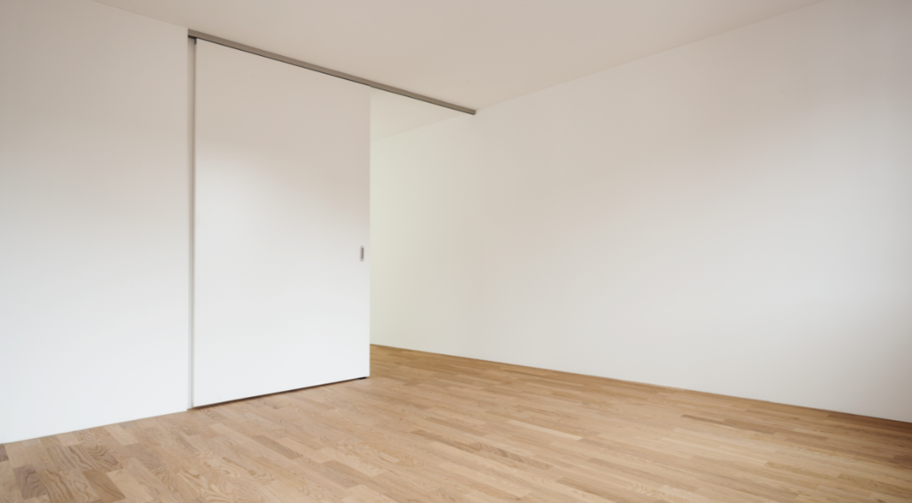 Ambiente sem mobílias ou decoração, com piso de madeira clara e paredes claras, com portas que se camuflam.