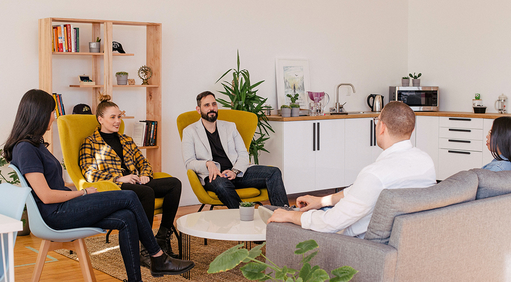 Espaço de descanso de um coworking, com decoração de estantes, livros e plantas. Nele, também há um sofá com pessoas sentadas conversando.