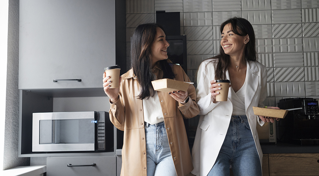 Copa de um espaço de coworking, com revestimento na parede, móveis planejados, decoração minimalista e duas mulheres conversando em primeiro plano.