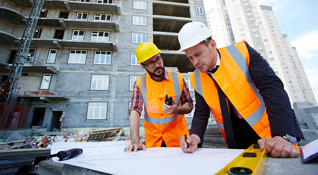 Homens com equipamento de proteção se debruçando em uma planta e fazendo gestão de obra, enquanto a obra está acontecendo atrás deles.