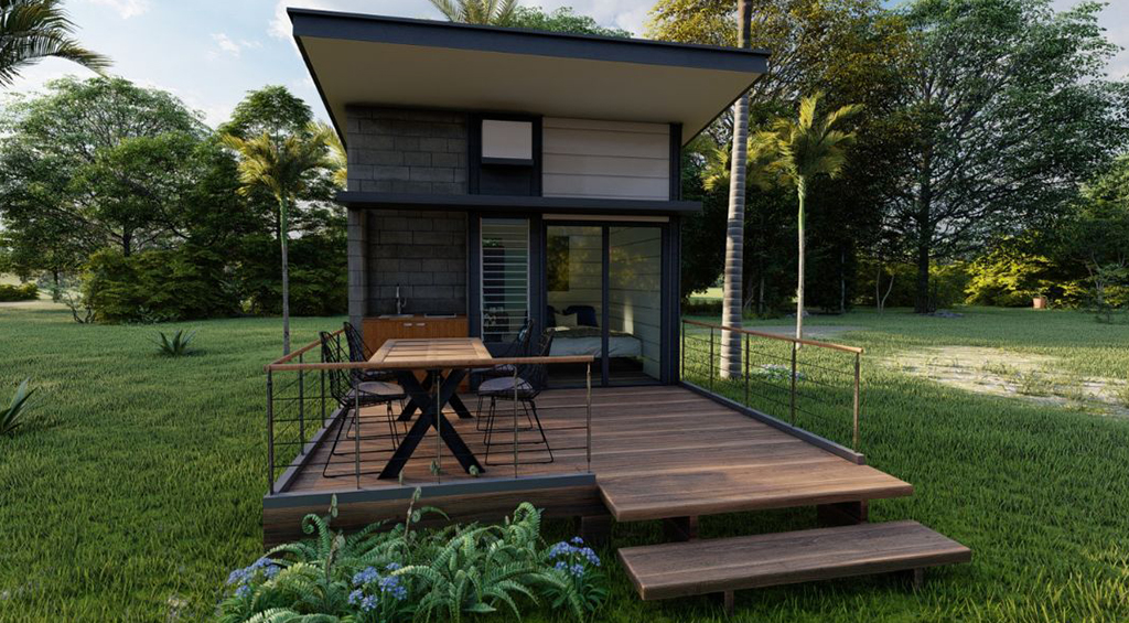 Tiny house fixa, com deck de madeira na área externa, em um extenso gramado com árvores atrás.