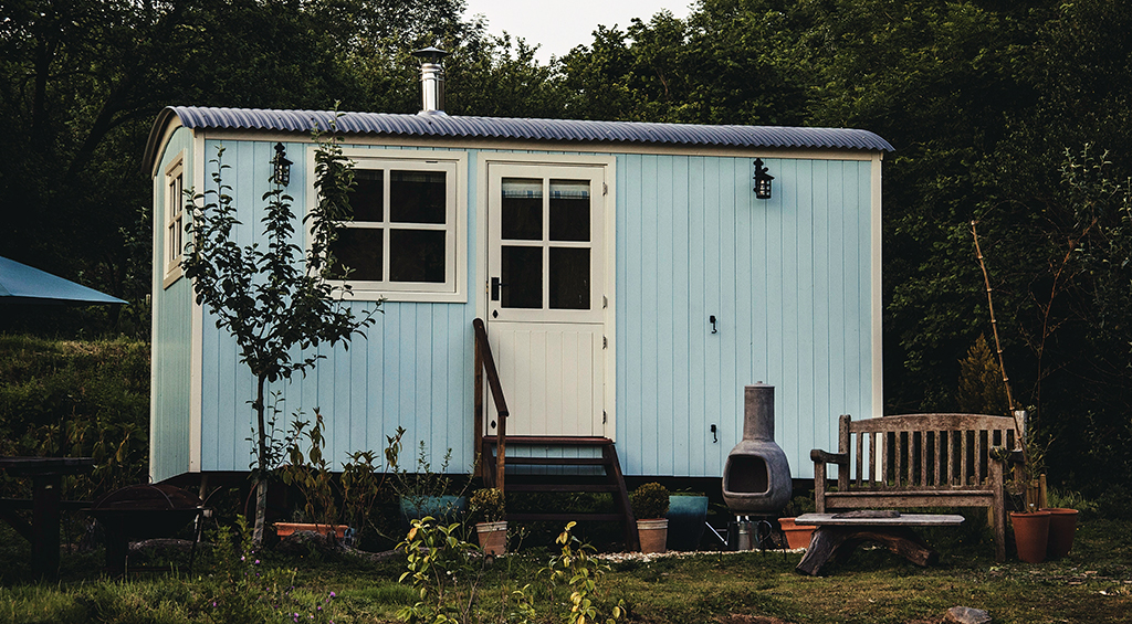 Tiny house transportável de madeira pintada de azul claro, em um gramado e com árvores atrás.